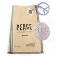 braune Papiertüte mit drei grauen Streifen links und rechts oben einem grauen Anhänger mit Peace Zeichen, auf der Tüte in Schwarz die Beschriftung "PEACE". Auf der Tüte liegt eine runde graue Seife mit Peace Zeichen