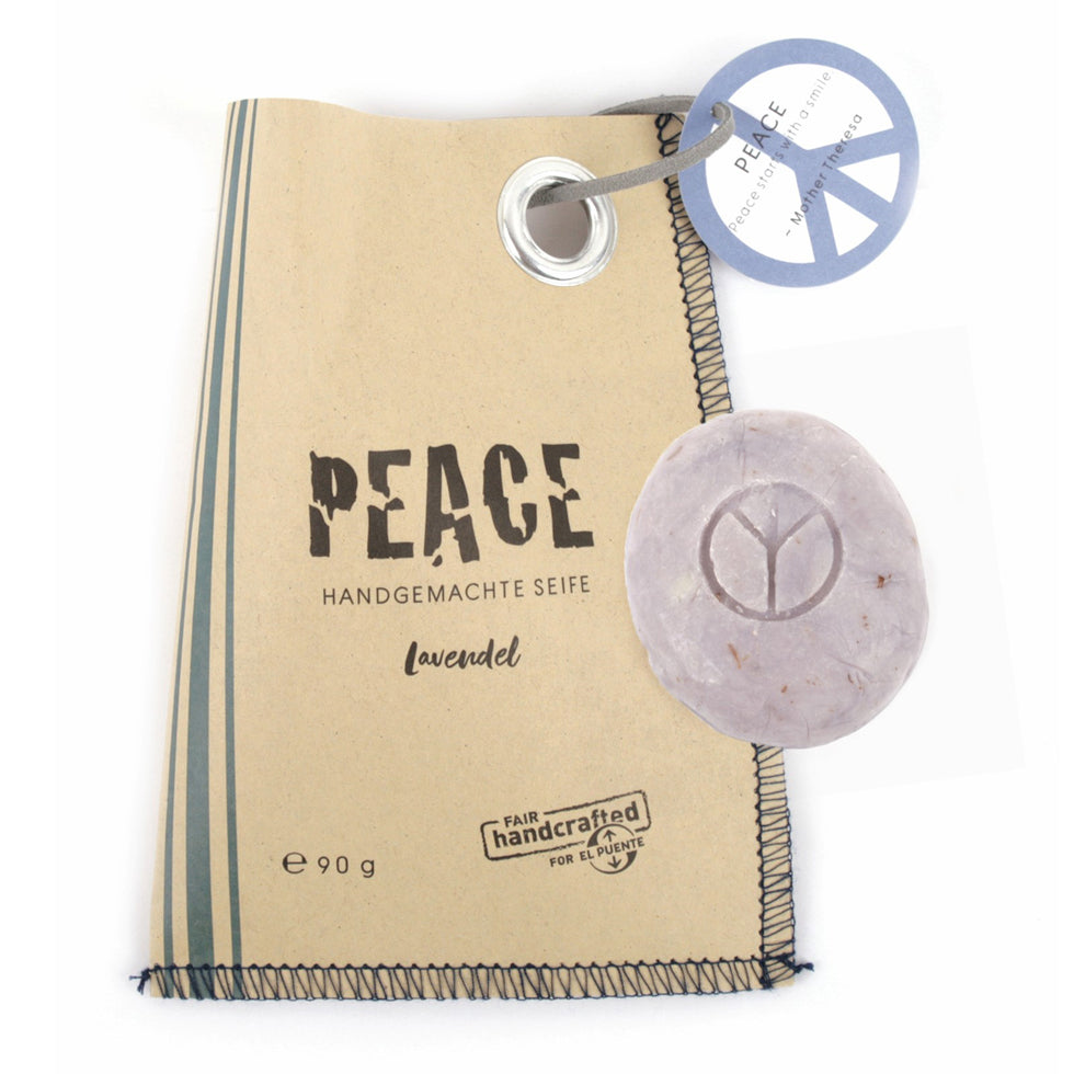 braune Papiertüte mit drei grauen Streifen links und rechts oben einem grauen Anhänger mit Peace Zeichen, auf der Tüte in Schwarz die Beschriftung "PEACE". Auf der Tüte liegt eine runde graue Seife mit Peace Zeichen