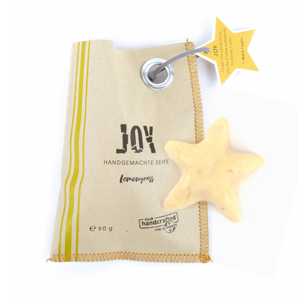 Braune Papiertüte, links mit drei gelben Streifen, mittig die Aufschrift JOY in Schwarz. Oben rechts befindet sich ein gelber Sternanhänger. Auf der Tüte liegt ein Stück gelbe Seife in Sternform