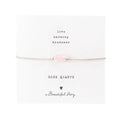Zartes Armband: silberfarbenes Baumwollband mit einem Rosenquarz-Edelstein auf einer Grußkarte. Oben stehen die Worte "Love, Harmony, Kindness", unten ein Rosenquarz, ein Herz und "A Beautiful Story".