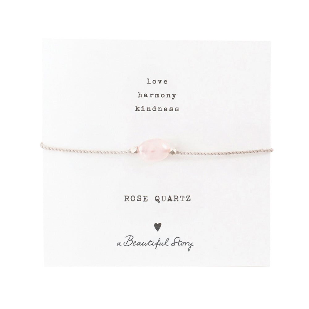 Zartes Armband: silberfarbenes Baumwollband mit einem Rosenquarz-Edelstein auf einer Grußkarte. Oben stehen die Worte "Love, Harmony, Kindness", unten ein Rosenquarz, ein Herz und "A Beautiful Story".
