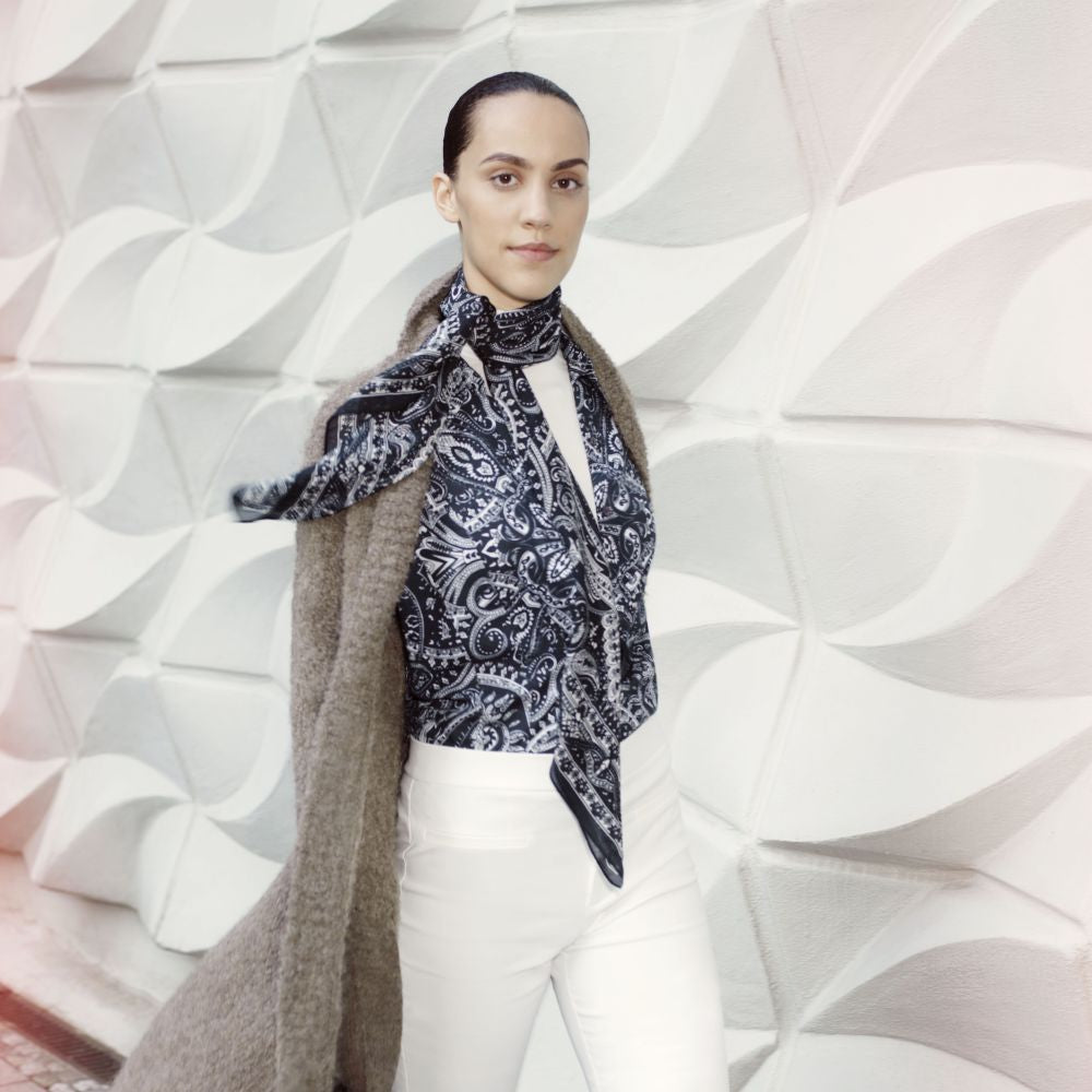 young woman wears a black blouse with a white print, white trousers and an open brown knitted coat