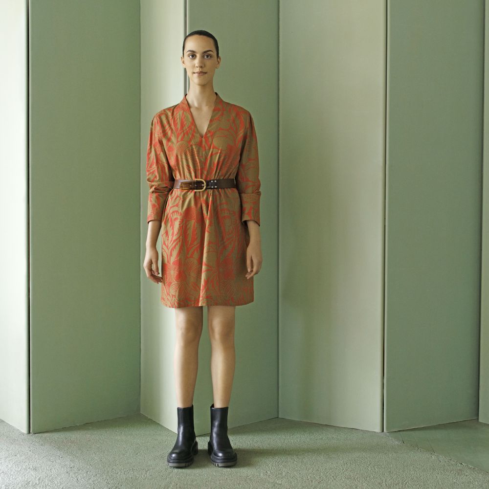 junge Frau in einem kurzen braunen Kleid mit orangefarbenem Muster, sie trägt einen braunen Gürtel um die Tailie sowie schwarze Kurzstiefel