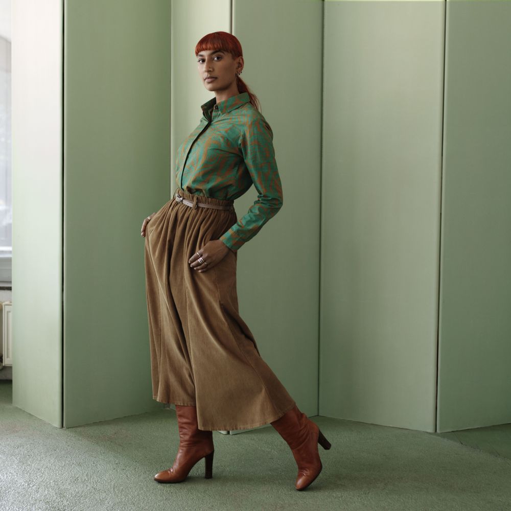 Young lady in brown trouser skirt and brown boots, wearing a dark green blouse with pattern