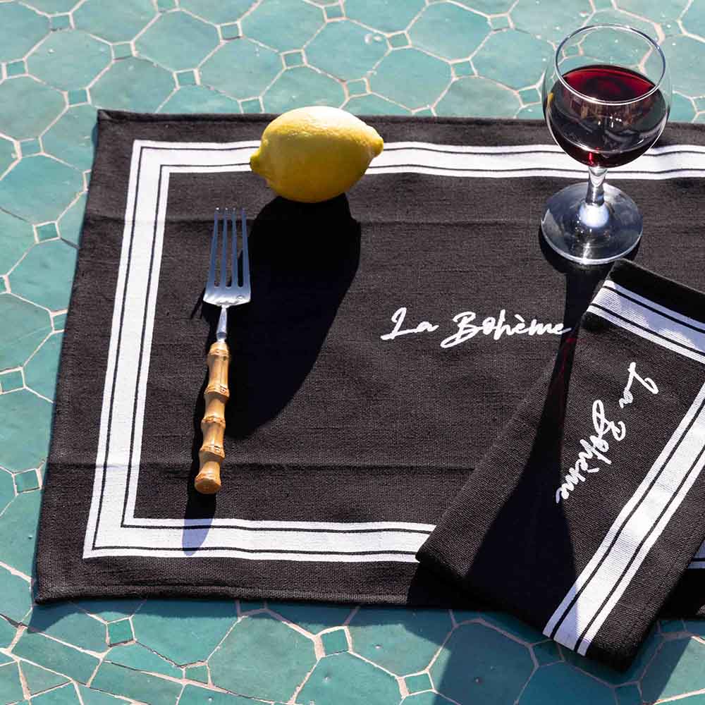 schwarz-weißes Platzset mit weißer Schrift auf einer Tischdecke, auf dem Set liegt eine passende Serviette, eine Gabel, eine Zitrone und ein Glas Rotwein