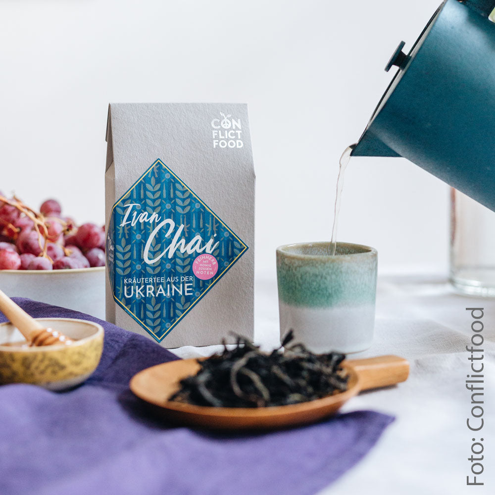 In the middle you can see a grey packet of tea with a blue label, in the background a bowl of grapes. In front of the tea is a small bowl of honey and the tea in a wooden bowl. Next to the tea, a teacup in which water is poured from a kettle