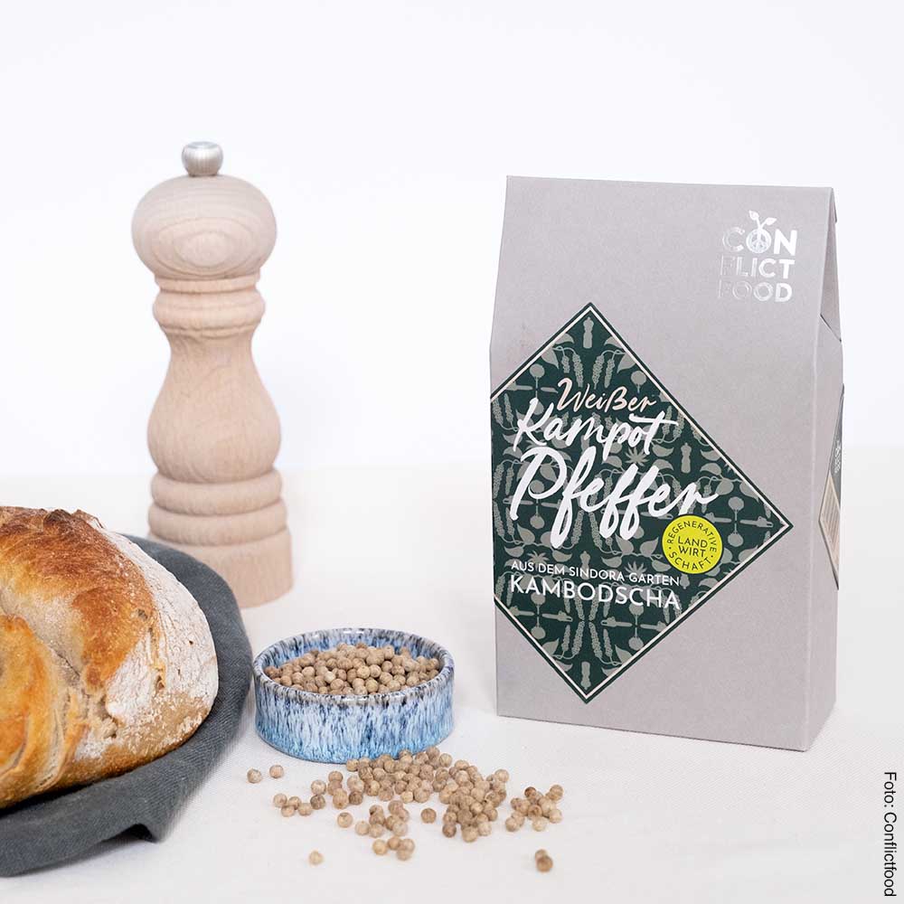 links liegt ein Laib Brot, dahinter eine Pfeffermühle aus Holz, davoor ein Schälchen mit Pfefferkörnern neben einer grauen Papiertüte mit mittigem Etikett