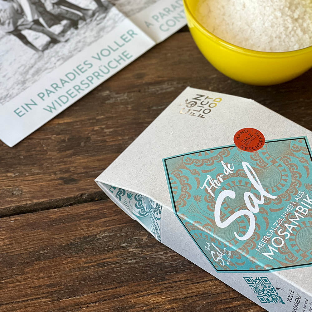 In the foreground:  A box of Fleur de Sel lies on a wooden table, next to a yellow bowl of salt flakes.