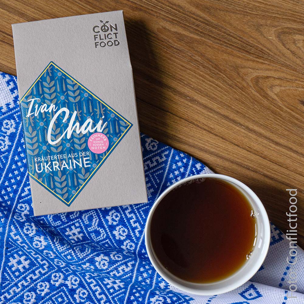 grey tea bag with a blue label in the middle lies next to a cup of tea on a blue and white blanket on a wooden table