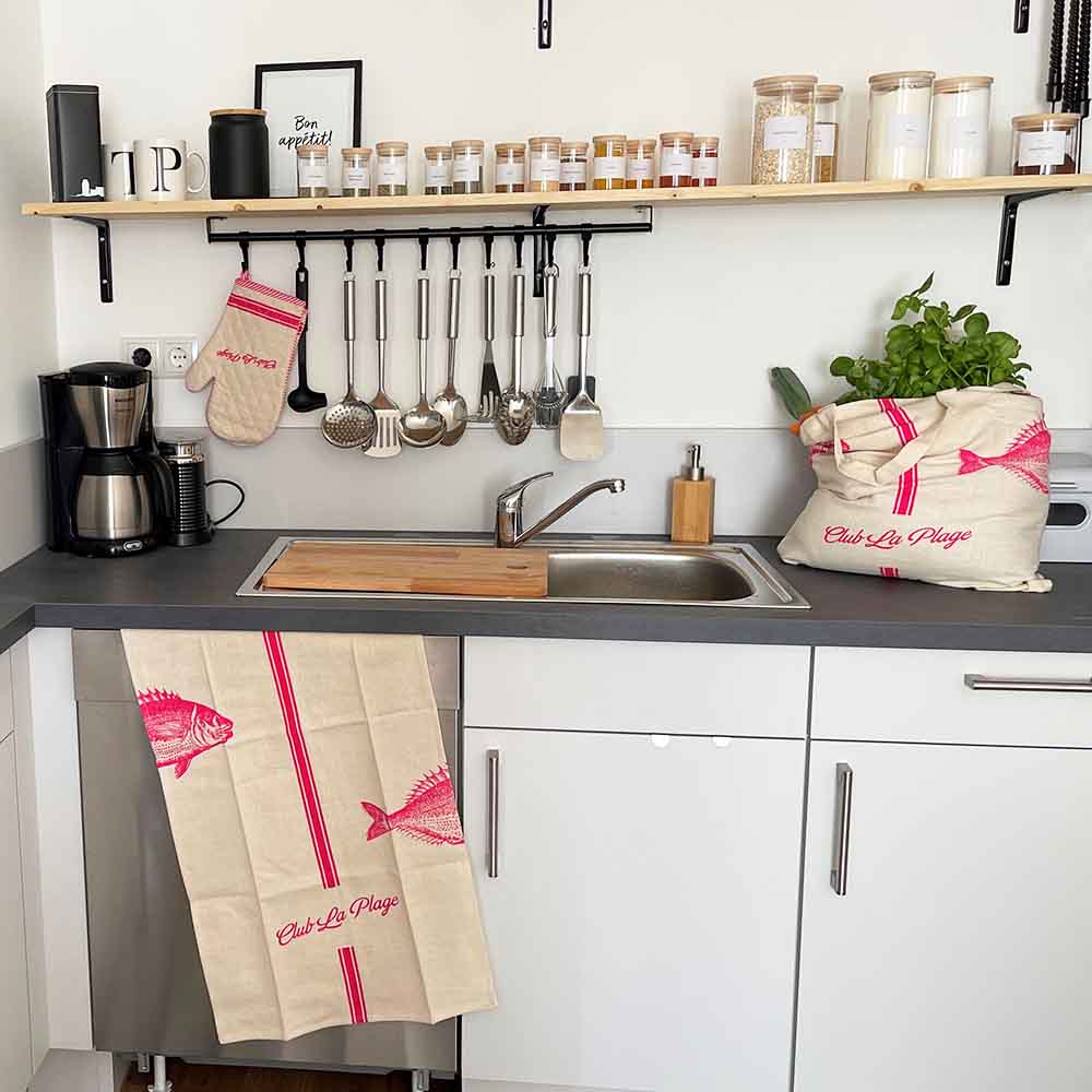 Küchenzeile mit Küchenutensillien am Haken über der Spüle, einem Regal darüber mit Gewürzen, eine hängende pink- weiße Schürze, Kochhandschuhe sowei eine gefüllte Einkaufstasche in der gleichen Farbe