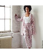 junge Frau mit rosa beruckter Pyjamahose, passendem, geöffnetem Kimono, darunter trägt die Dame ein weißes Tank-Top. Die Frau steht angelehnt an einer weißen Wand
