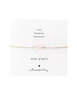 Zartes Armband: silberfarbenes Baumwollband mit einem Rosenquarz-Edelstein auf einer Grußkarte. Oben stehen die Worte "Love, Harmony, Kindness", unten ein Rosenquarz, ein Herz und "A Beautiful Story".