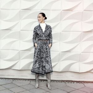 junge Frau in trägt einen schwarzen Rock mit weißem Print und einer gleichen Bluse. Dazu trägt sie einen braunen Ledergürtel, graue Stiefel, unter der Bluse ein weißes Shirt. 