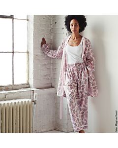 junge Frau mit rosa beruckter Pyjamahose, passendem, geöffnetem Kimono, darunter trägt die Dame ein weißes Tank-Top. Die Frau steht angelehnt an einer weißen Wand