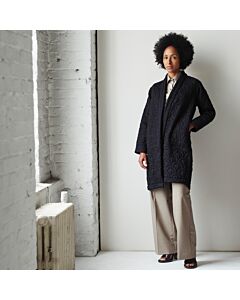 Junge Frau mit schwarzem Haar trägt einen schwarzen Steppmantel, eine graue Hose und schwarze Sandalen