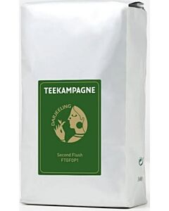 Tea packaging, silver bag with green label. The label features a round, gold-coloured logo. The logo consists of the word Darjeeling and shows a person in profile holding a tea leaf in their hand and holding it to their nose.